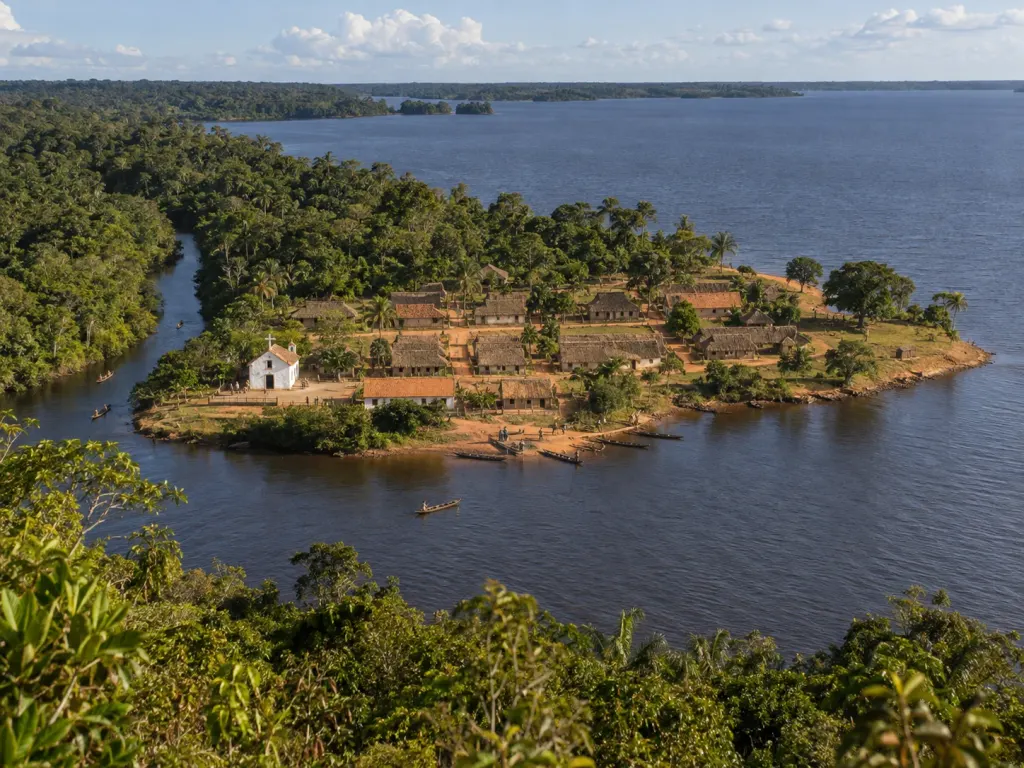 Vista do alto de um morro de uma pequena vila numa península baixa, com a igreja caiada, casas de palha em quadras sobre terra vermelha, canoas no porto e um igarapé entrando na floresta