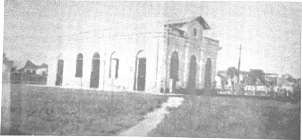 Fotografia antiga em preto e branco, com retícula de impressão, do prédio térreo do antigo mercado com portas em arco, diante de um gramado