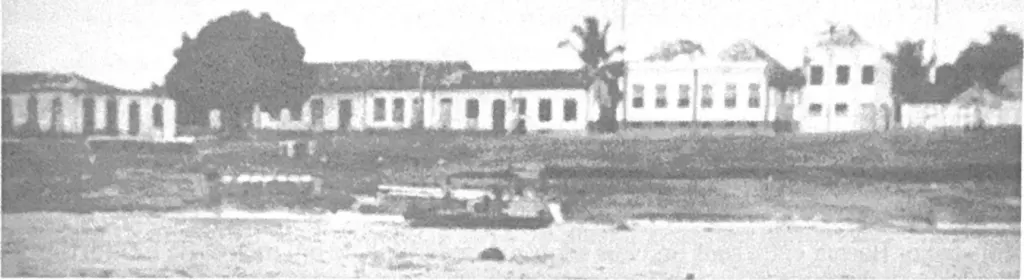 Fotografia antiga em preto e branco, com retícula de impressão, da frente da cidade vista do lago: casas térreas e sobrados sobre o barranco, uma árvore grande, uma palmeira e um barco na água