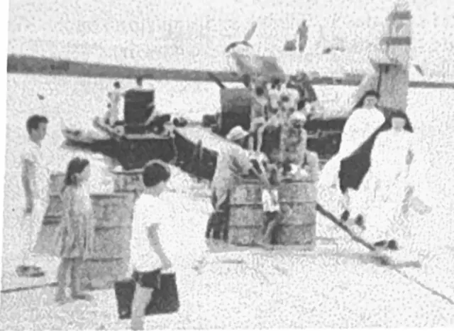 Fotografia antiga em preto e branco, com retícula de impressão, da balsa com o hidroavião ao fundo, tambores, crianças e duas freiras