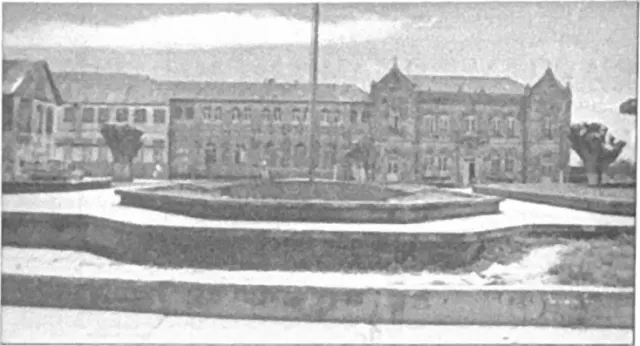 Fotografia antiga em preto e branco, com retícula de impressão, da praça com canteiros octogonais e mastro diante do Palácio Episcopal e do Seminário