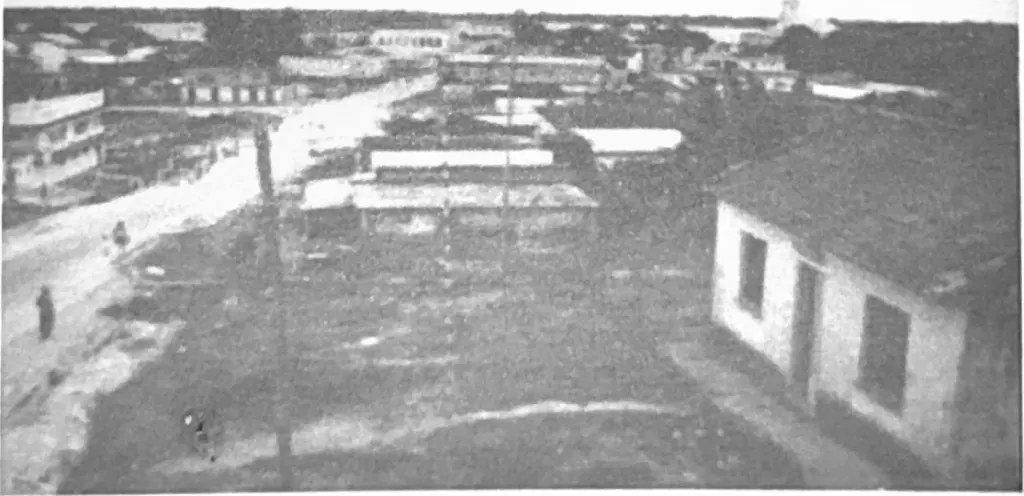 Fotografia antiga em preto e branco, com retícula de impressão, de uma rua larga de terra vista do alto, com poste e casas