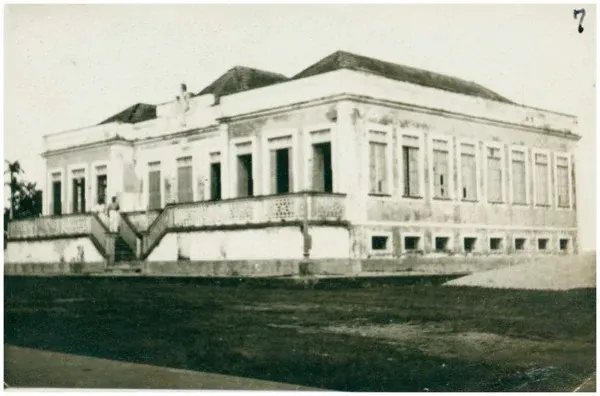 Fotografia antiga em preto e branco de um prédio térreo largo com escadaria dupla e muitas janelas, diante de um gramado