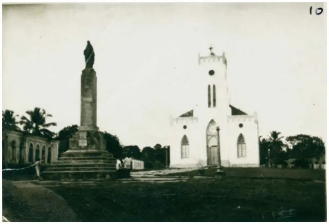 Fotografia antiga em preto e branco da praça com um monumento alto sobre pedestal e a igreja de torre única com janelas ogivais